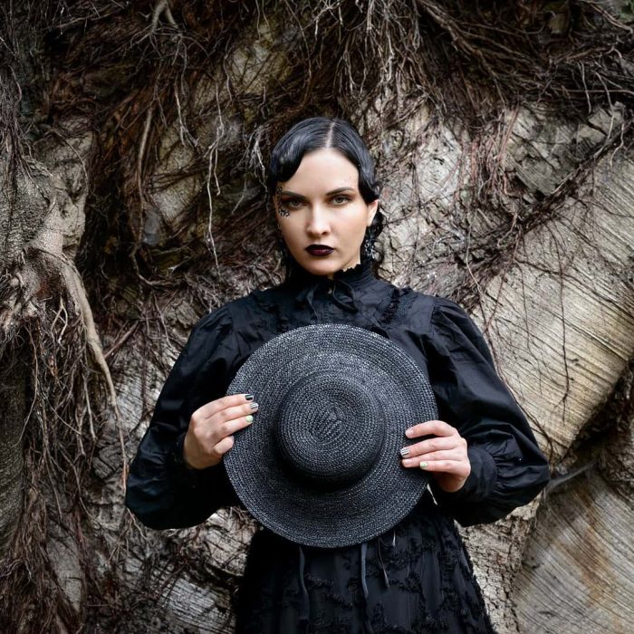 Fashion portrait of a woman wearing a black hat and outfit posing in front of a tree