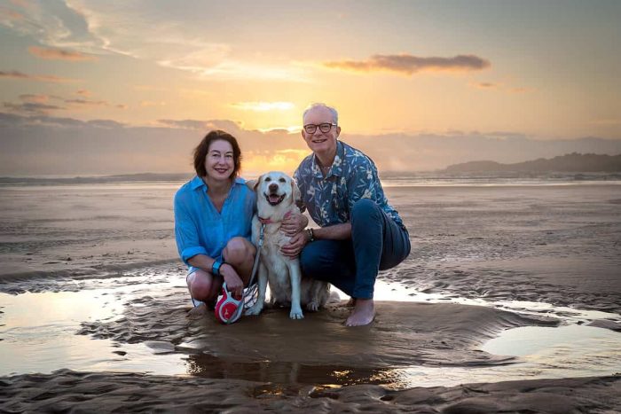 Couple kneeling beside their dog on Boambee Beach at sunset, smiling, lifestyle pet portrait photography