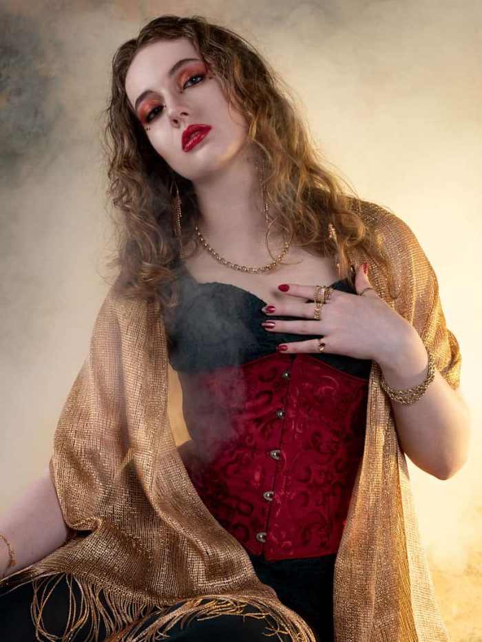 Studio portrait of woman posing with gold smoky background, wearing red, gold and black outfit with bold makeup