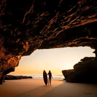 Lady with surfboard standing in a cave entrance looking out at the water at sunrise
