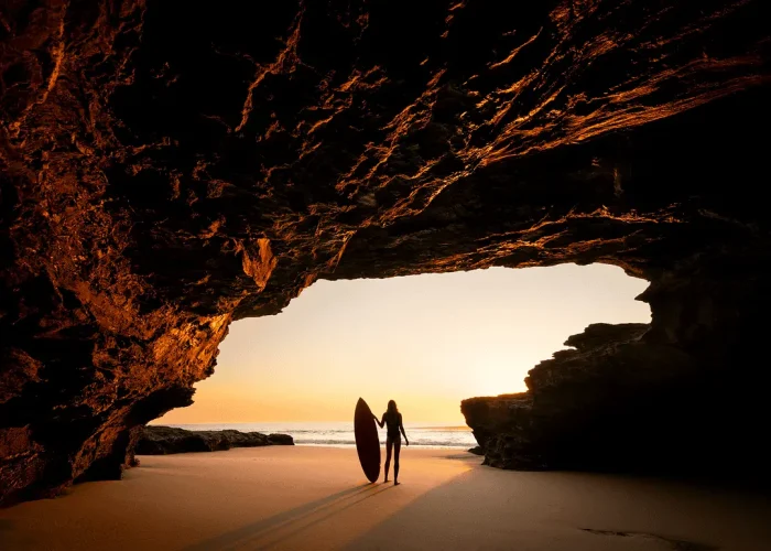 Lady with surfboard standing in a cave entrance looking out at the water at sunrise